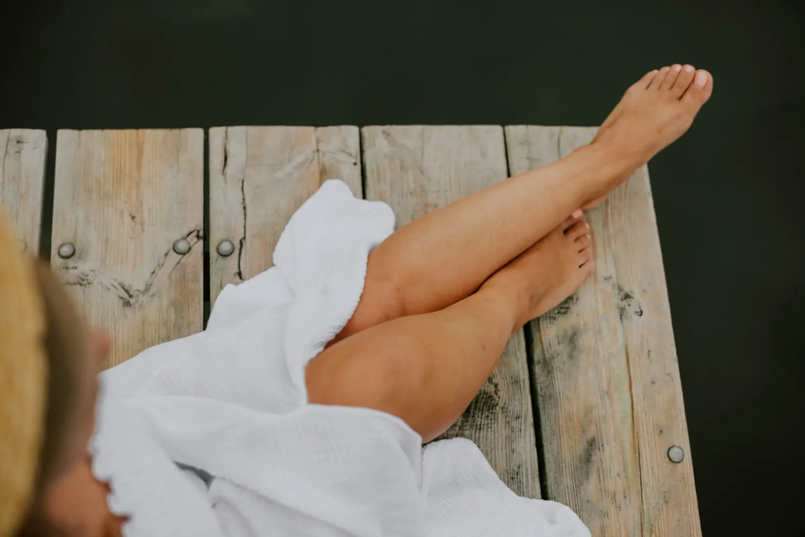 Smooth bare legs crossed on a wooden dock beside a white towel